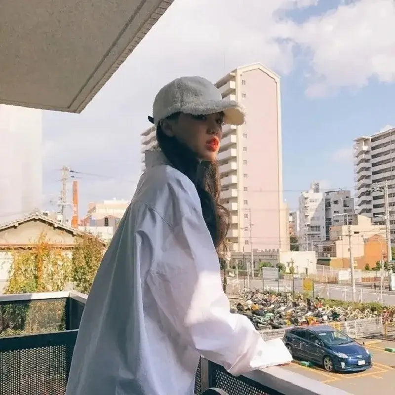 Woman wearing a chic oversized white shirt and cap, posing on a balcony with urban scenery.