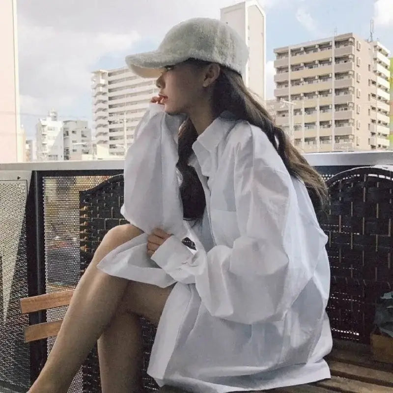 Woman in oversized white shirt and cap, sitting casually with urban backdrop, showcasing minimalist chic style.