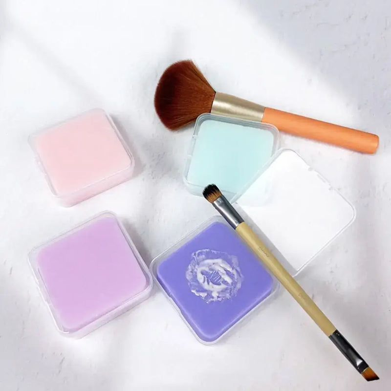 Assorted makeup brush cleaner soaps in storage containers with brushes on a white background.