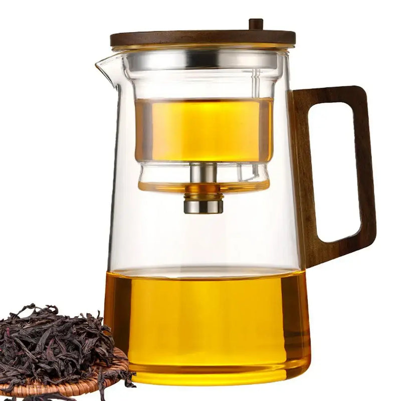 Glass teapot with removable infuser, wooden lid, and tea leaves beside it, ideal for brewing tea.