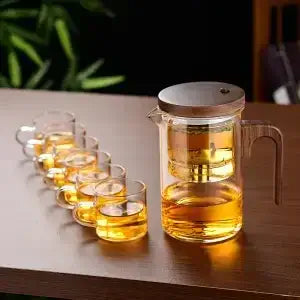 Heat-resistant glass teapot with stainless steel infuser and tea cups on a wooden table.