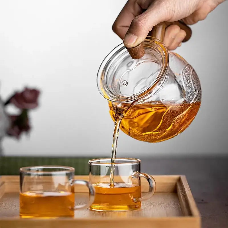 Pouring fragrant tea from a heat-resistant glass teapot into two clear tea cups on a wooden tray.