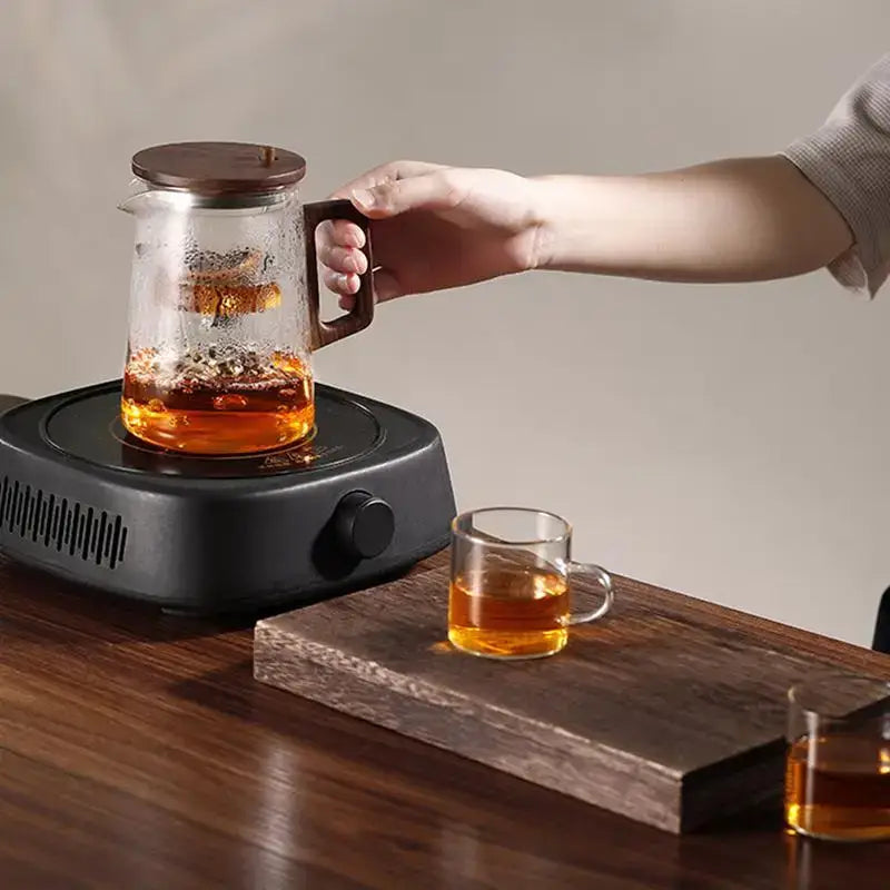 Glass teapot brewing tea on an electric stove with a wooden tray and tea cups.