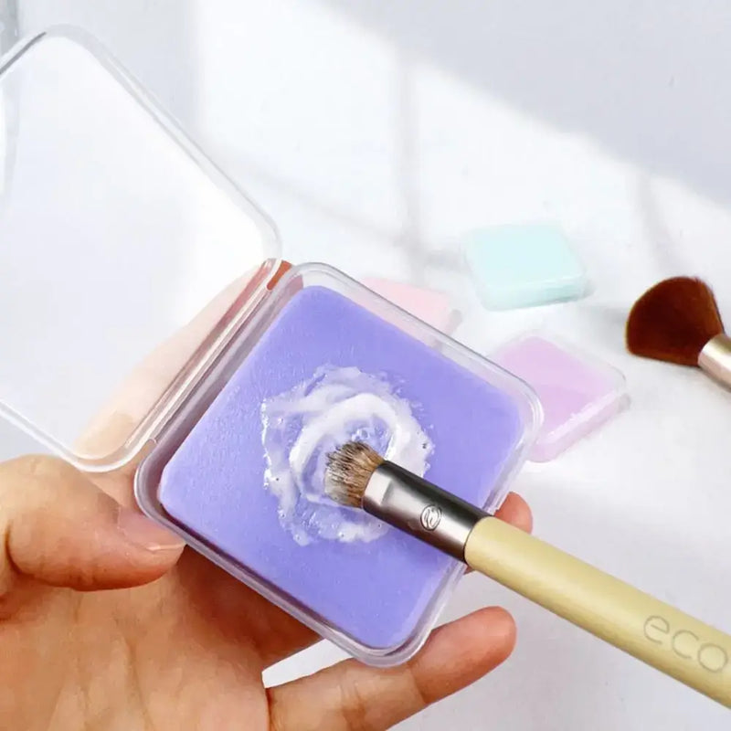 Hand holding a transparent storage box with purple makeup brush cleaner soap and a brush for cleaning.