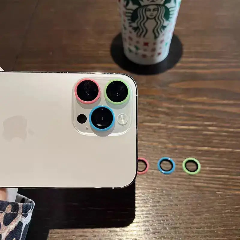 Colorful camera lens protectors on a smartphone, showcasing scratch-resistance and stylish design with a background of a coffee cup.