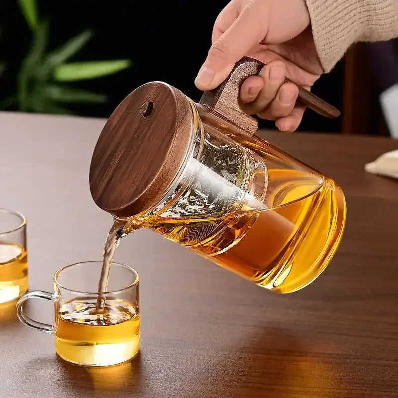 Heat-resistant glass teapot pouring tea into a cup, featuring stainless steel infuser and wooden lid.