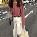 Woman in loose-fit long sleeve pink top, white pants, and gray sweater, walking down a street with parked cars.